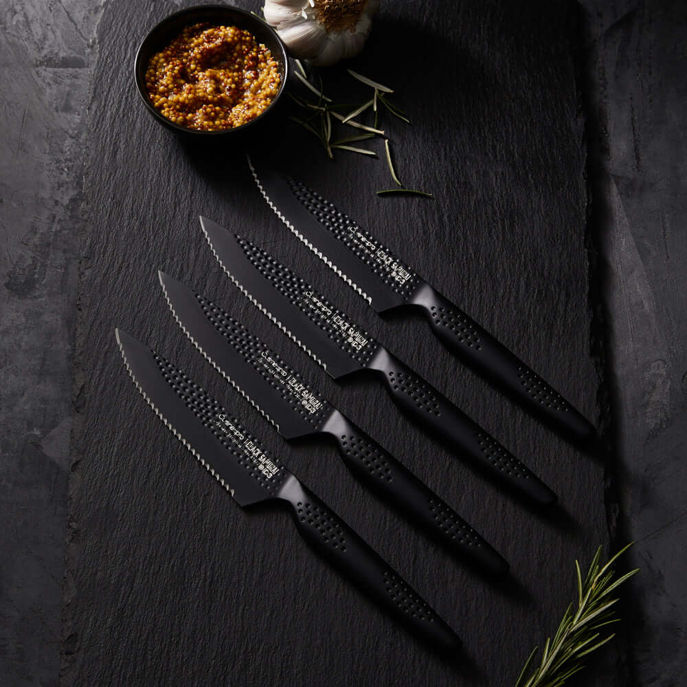 Cuisine::pro® iD3® BLACK SAMURAI™ 4 Piece Steak Knife Set displayed on a dark surface with a bowl of mustard and herbs.