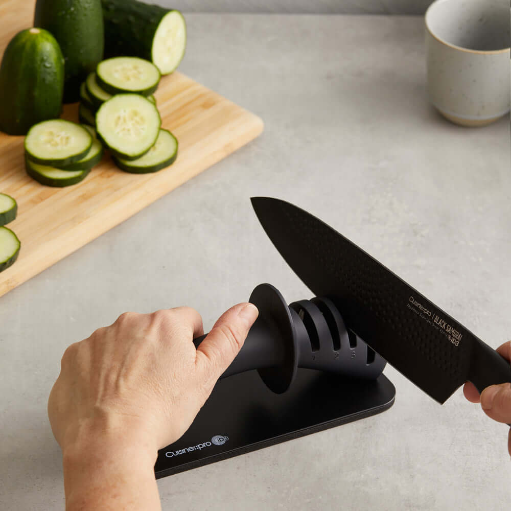 Using Cuisine::pro® iD3® BLACK SAMURAI™ 3-Step Knife Sharpener to sharpen a knife near a cutting board with sliced cucumbers.