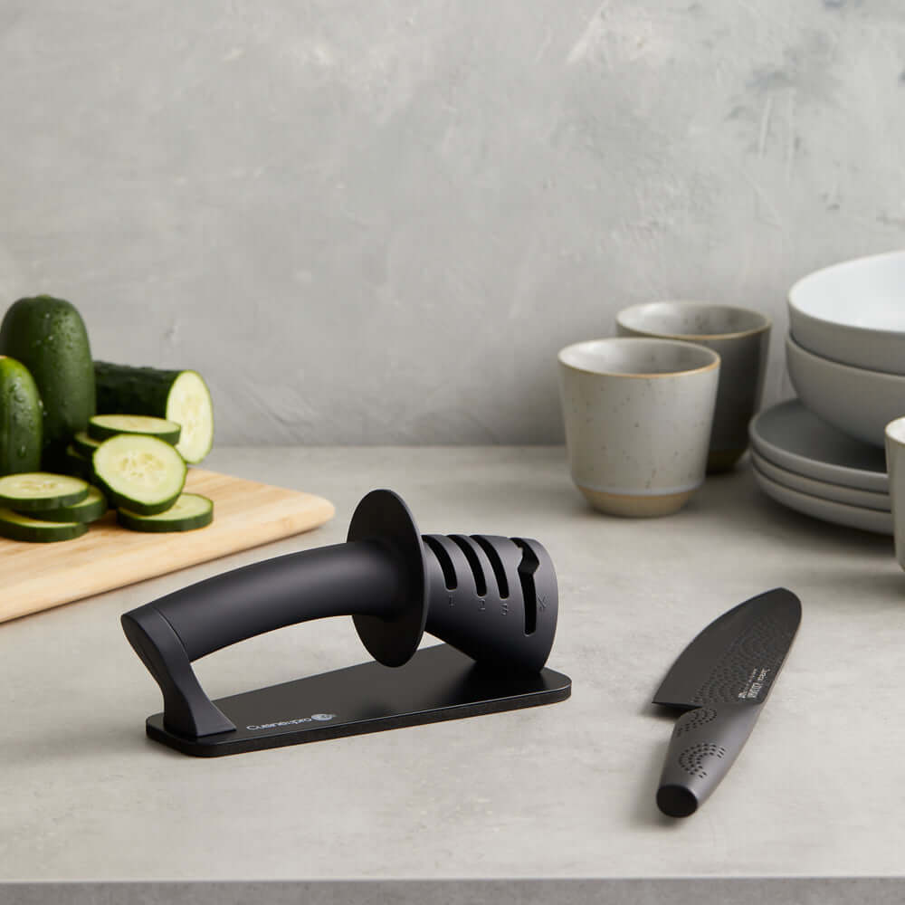 Cuisine::pro® iD3® BLACK SAMURAI™ 3-Step Knife Sharpener with knife on a kitchen counter beside sliced cucumbers and dishes